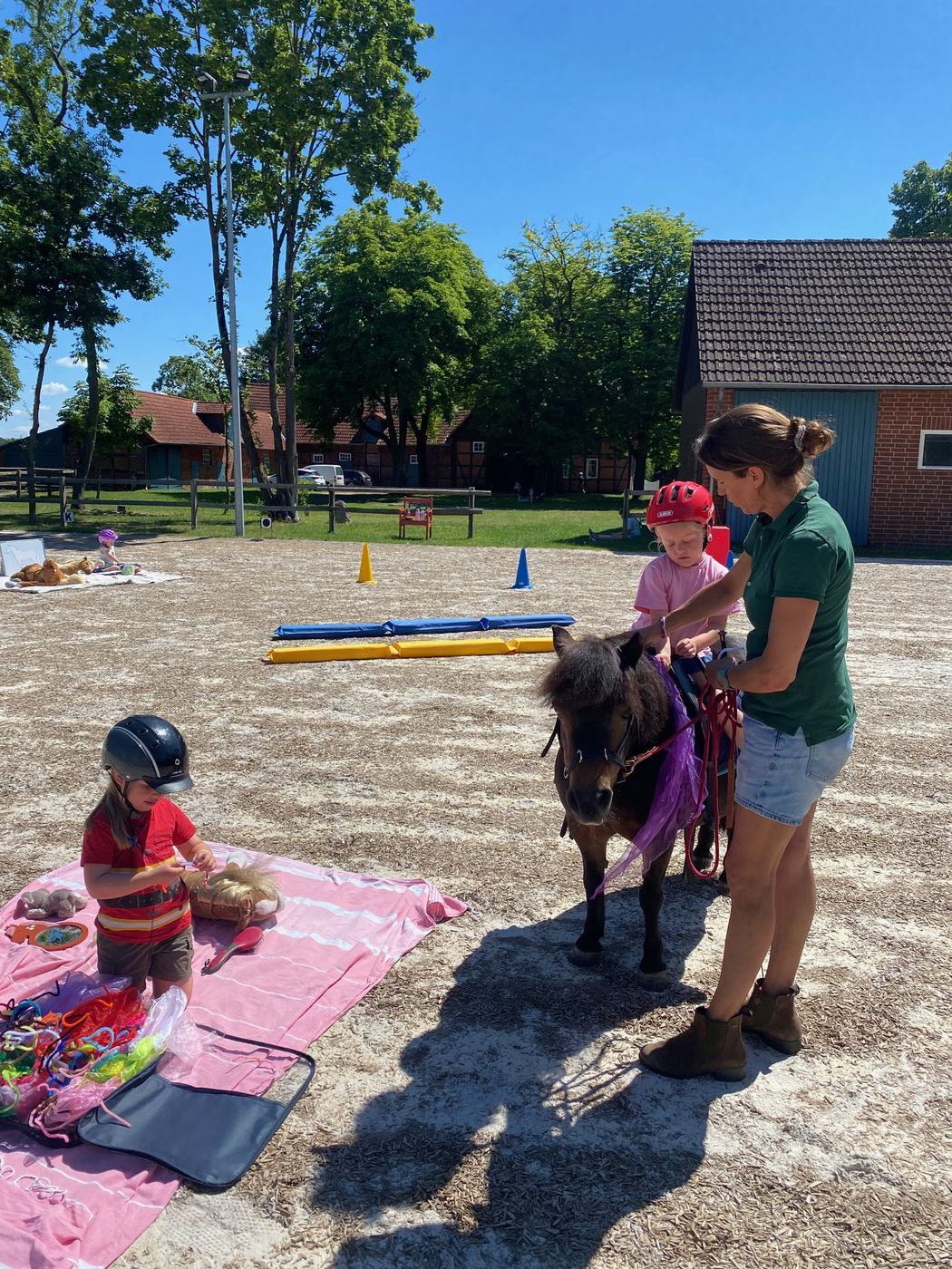 Kleines Pony mit lila Satteldecke auf dem Reitplatz, ein Kind spielt auf der Decke